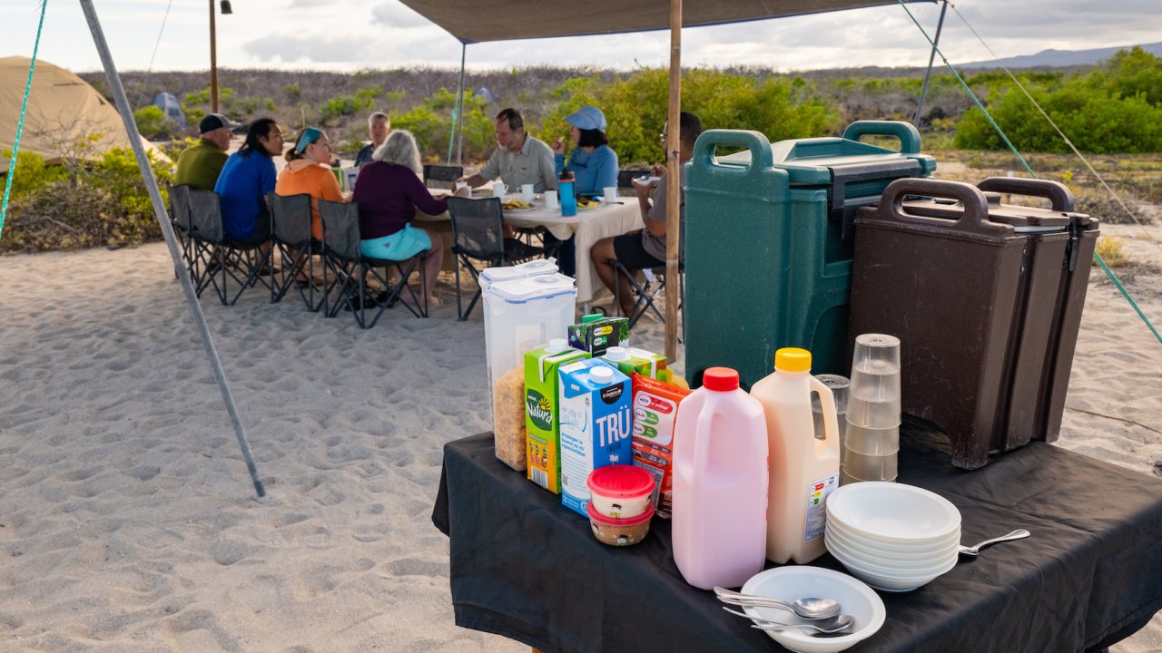 Appetizer table set up with milk, juice, water, cutlery, and bowls and plates at a campsite in the Galapagos