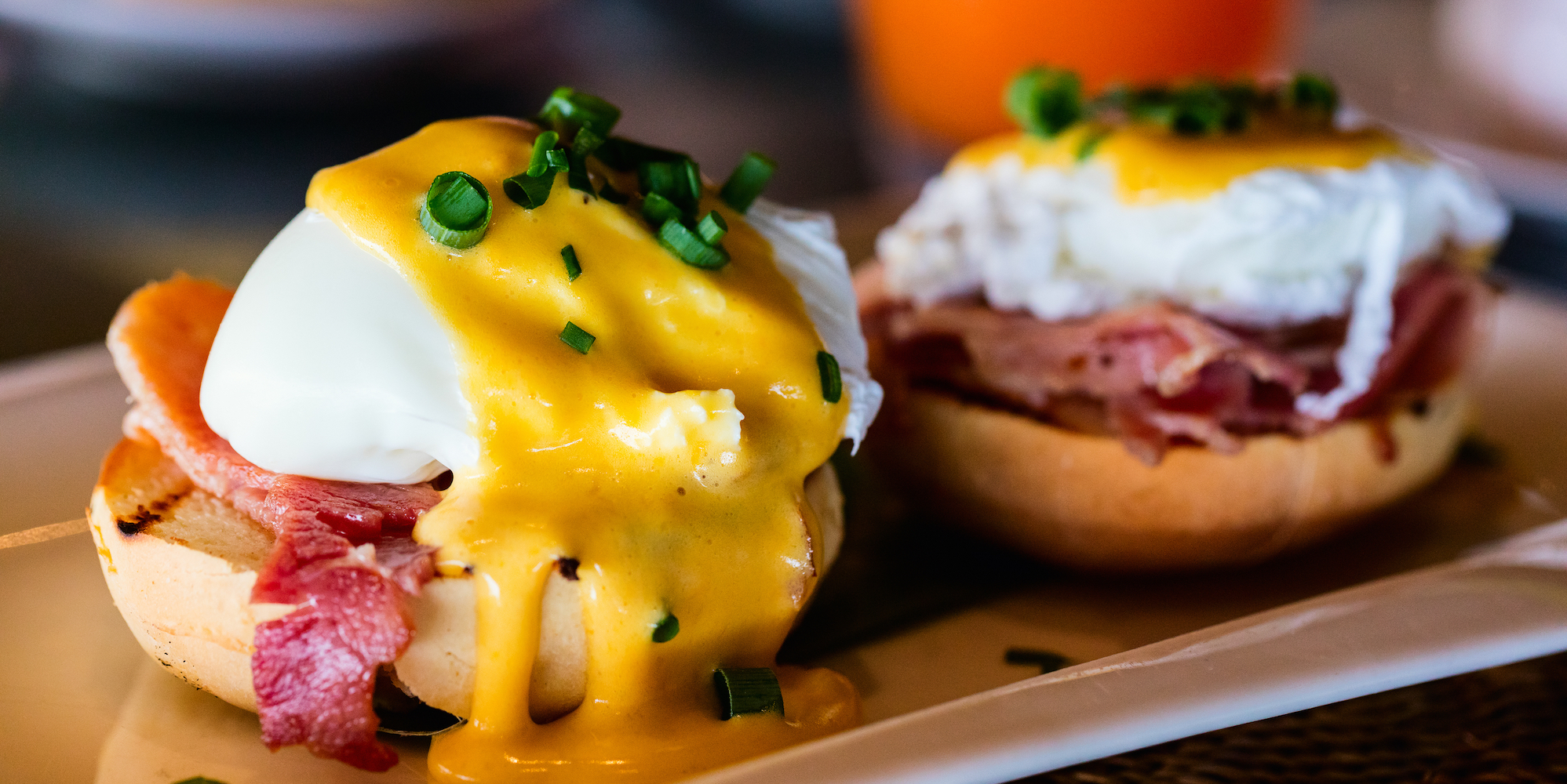 Close up on eggs Benedict side by side topped with scallions on a white rectangle serving plate 