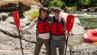 Two people wearing sunglasses, hats, and life jackets holding their paddles smile for a photo