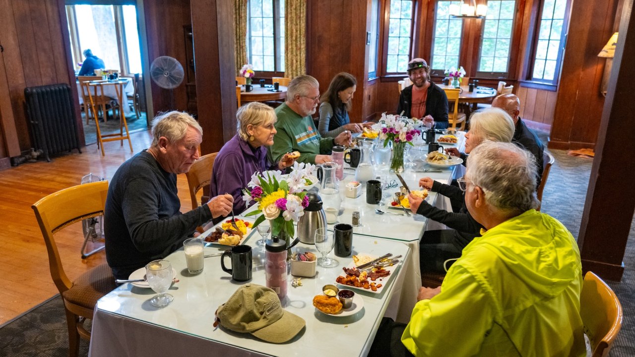 A group of older adults enjoying a meal together inside a cozy wood-paneled lodge, sharing stories and laughter after a day of adventure.