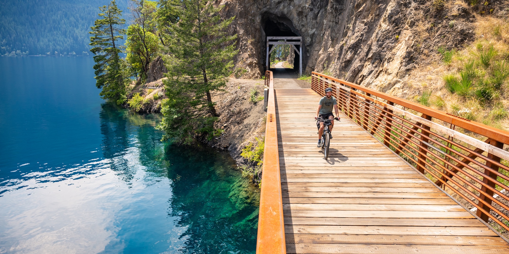 A man biking over a bridge crossing deep, clear, blue water in Olympic National Park at Lake Cresent.