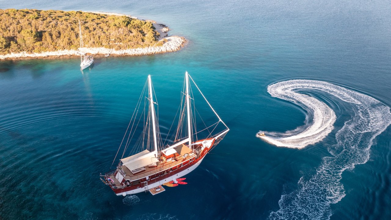 A sailboat anchored near a small island in clear blue water, with a jet ski circling nearby.
