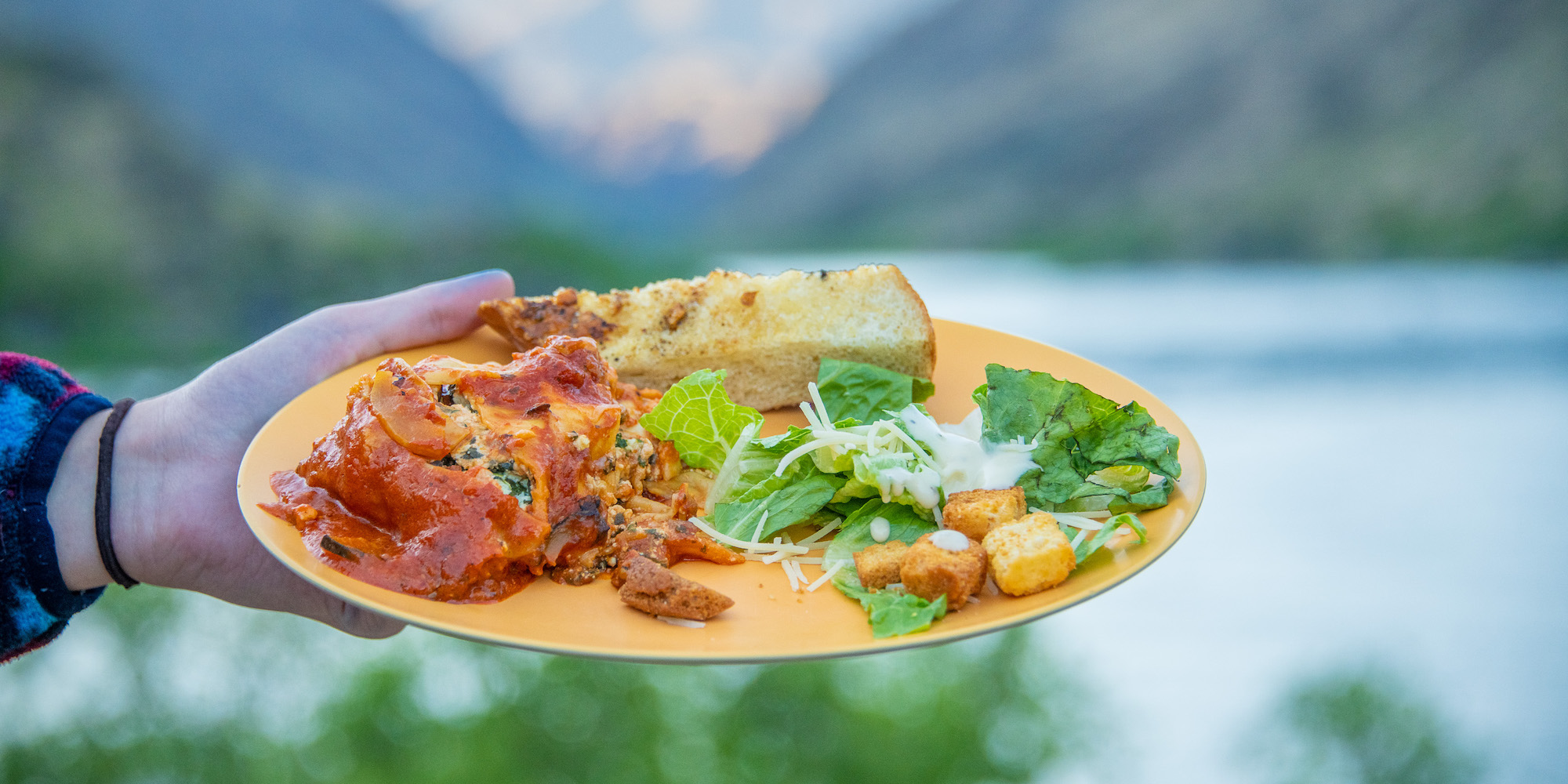 Someone holding a yellow plate of lasagna, caesar salad and garlic bread while camping