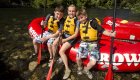 Three kids sitting on the edge of a red ROW Adventures raft on Idaho’s Clearwater River, smiling in yellow life jackets with paddles resting in the water.