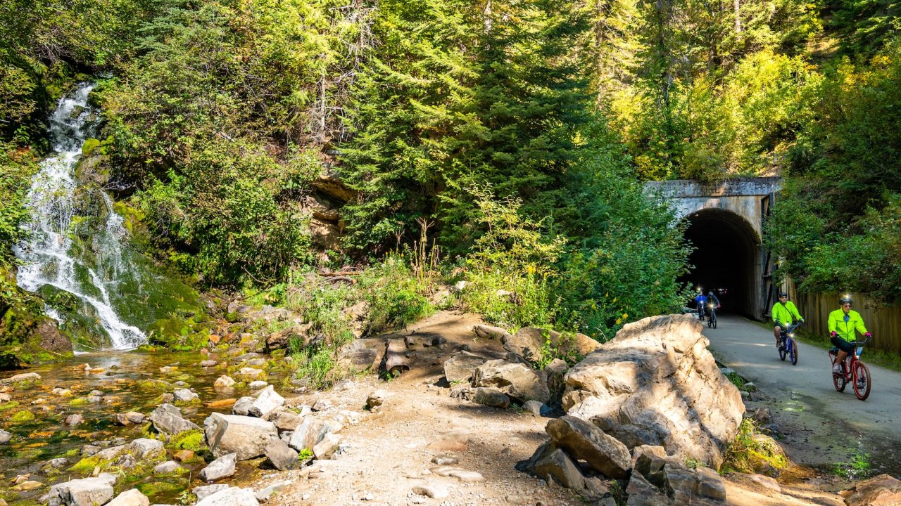 Group of cyclists ride past a waterfall and historic tunnel on the scenic Idaho Hiawatha Bike Trail.