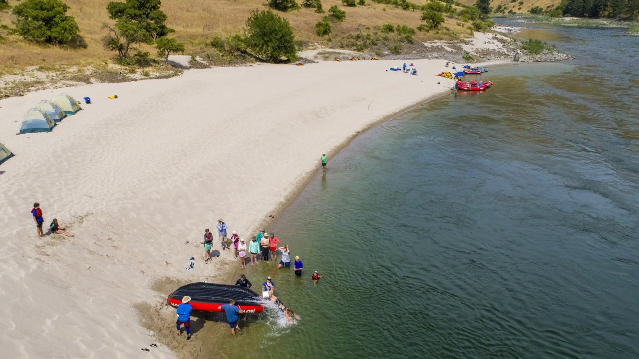 Families camping and playing on a sandy river beach with rafts nearby.