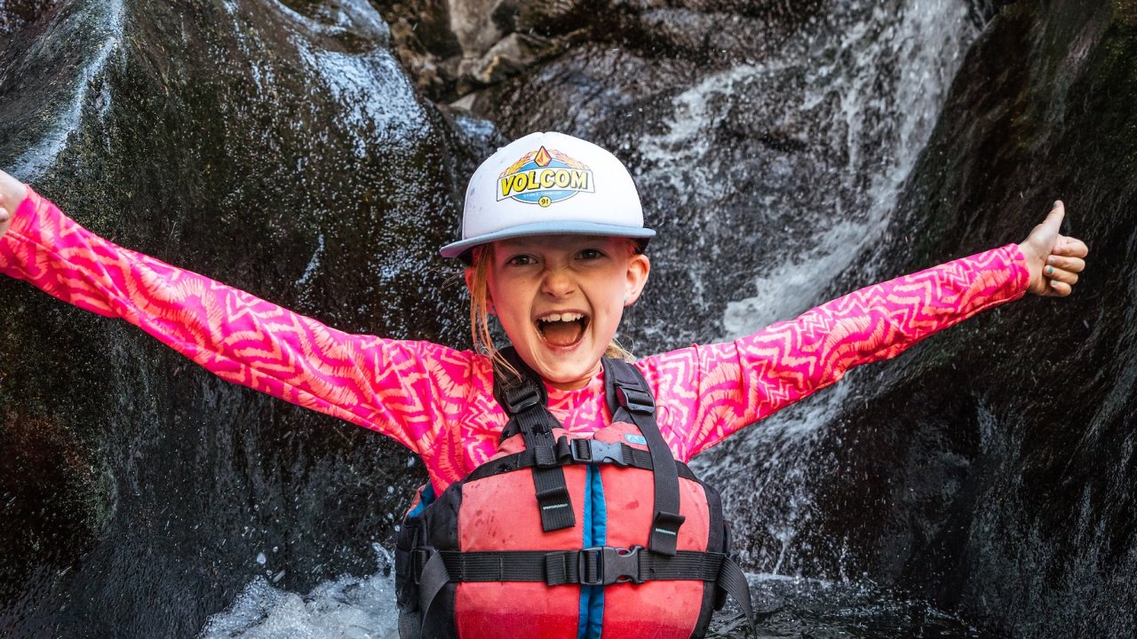 A young girl sitting at the base of a small waterfall and smiling while on a ROW Adventures family rafting trip in Idaho.