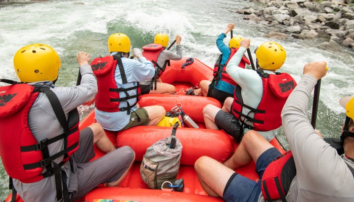 Picture from the back of a red paddle boat with six people in red PFDs and yellow helmets paddling through a rapid