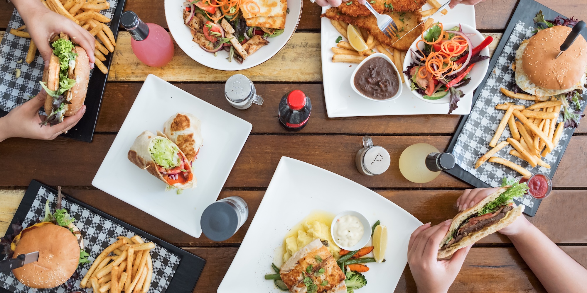 Food displayed out on a wooden picnic table as peoples hands hold the meal.