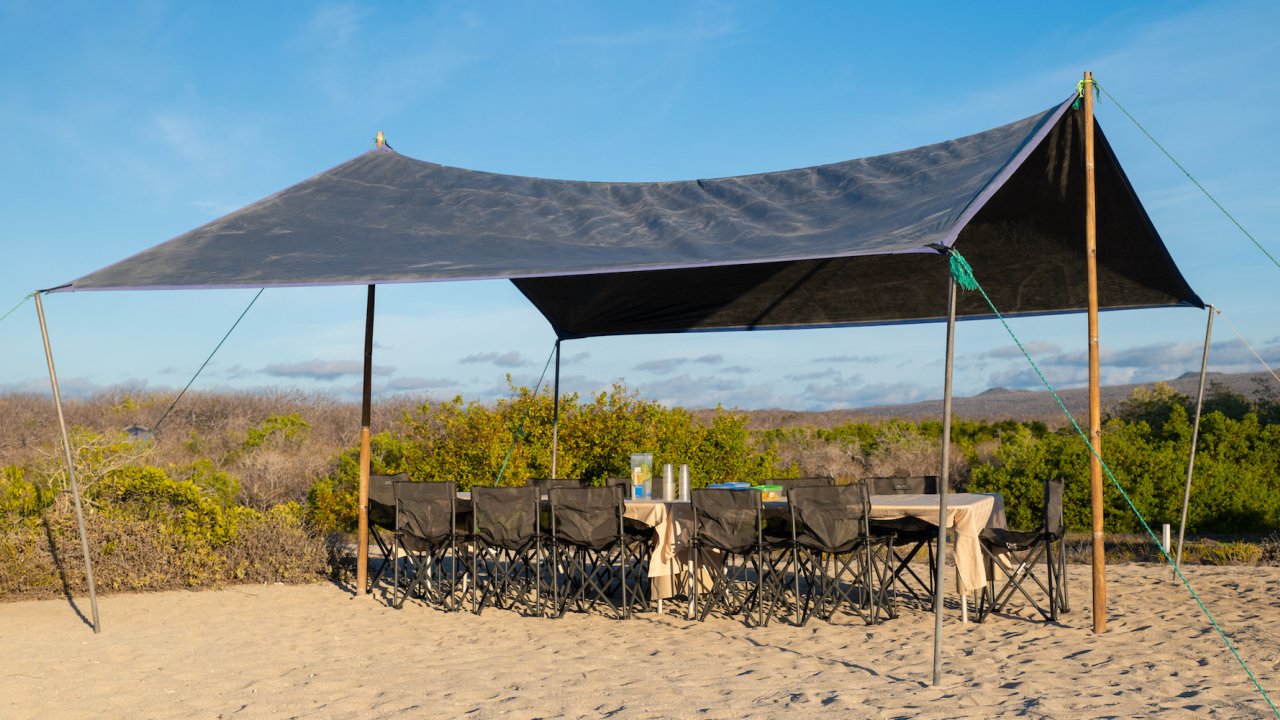 A big wind fly set up over a long table with camp chairs on a beach