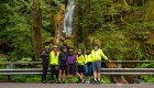 A group of cyclists in bright jackets posing in front of a waterfall in a mossy Pacific Northwest forest during a guided biking tour.