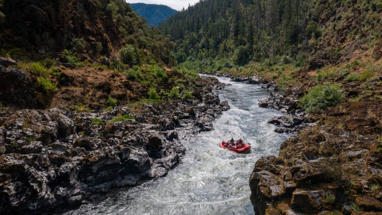 A red raft navigates whitewater rapids on the Rogue River in Oregon, surrounded by rugged canyon walls and forested hills, showcasing the thrill of Rogue River rafting adventures.