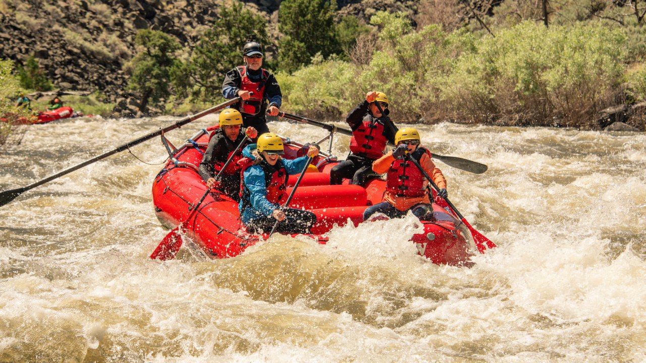 A man rowing a boat with two people sitting in front of him paddling and smiling through a rapid in Idaho