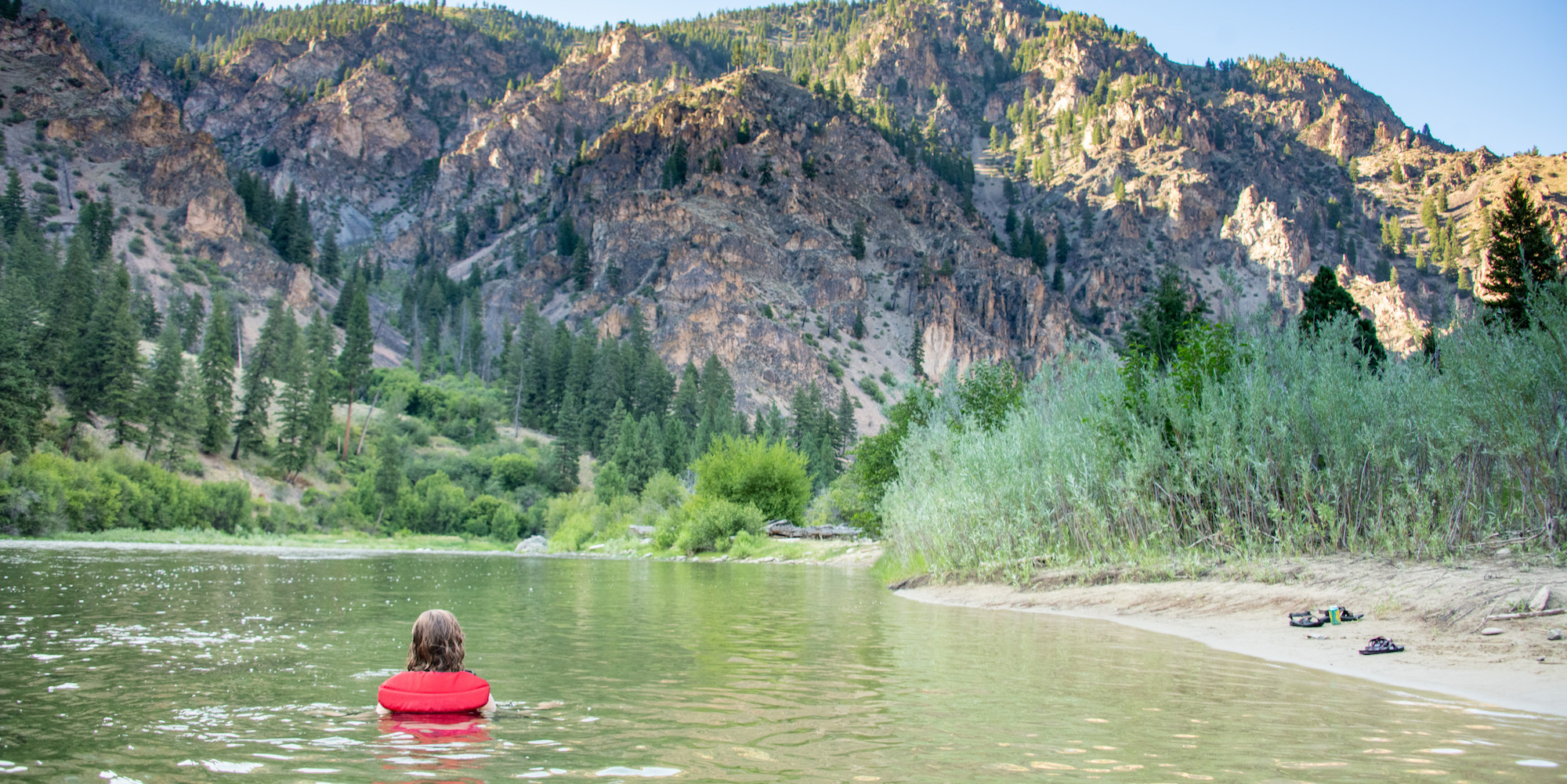 A girl in a red life jacket standing in the Salmon River looking at the mountains at sunset