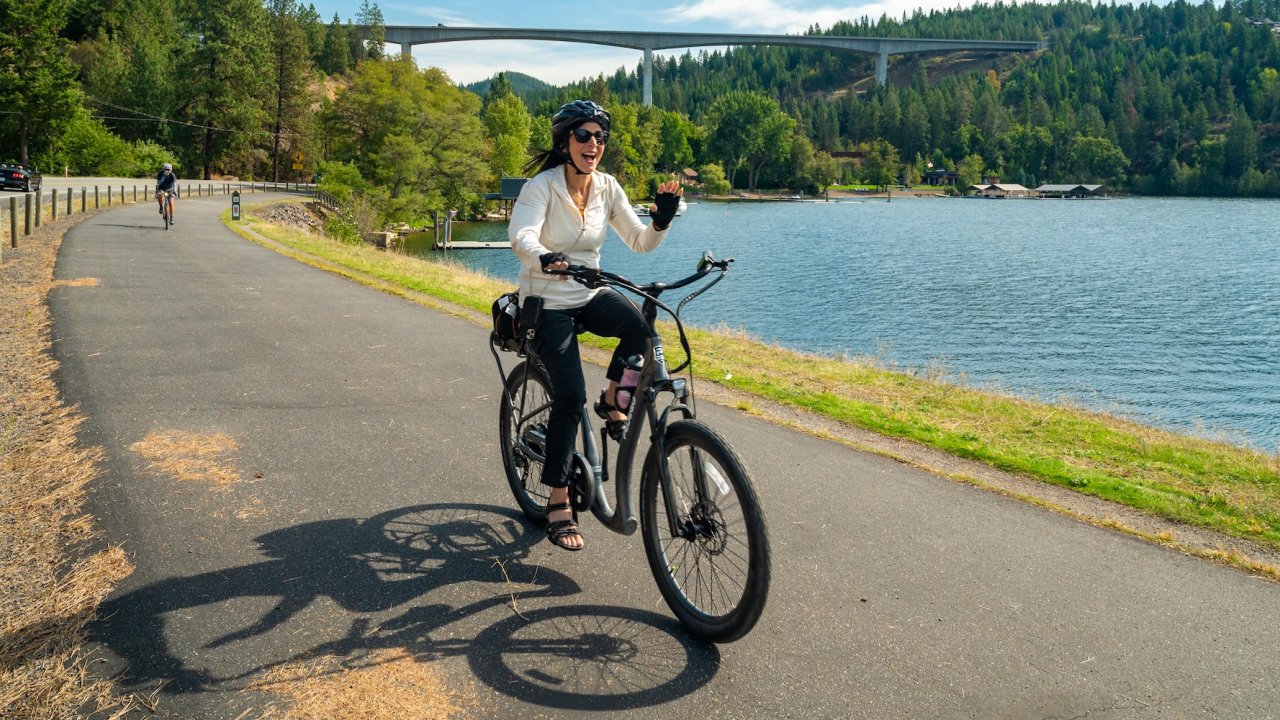 A smiling woman rides an e-bike along a scenic lakeside path in northern Idaho, with a large bridge and forested hills in the background.