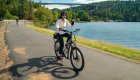 A smiling woman rides an e-bike along a scenic lakeside path in northern Idaho, with a large bridge and forested hills in the background.