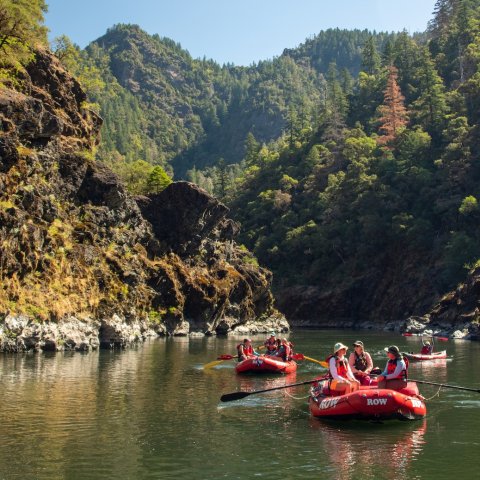 A couple of red ROW rafts floating down a calm section of the Rogue river in Oregon with rocky gorge walls surrounding them. 
