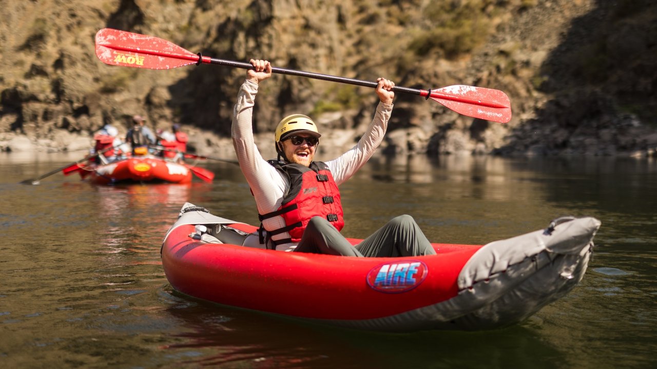 Man paddling an inflatable kayak on calm river, raising paddle in celebration.