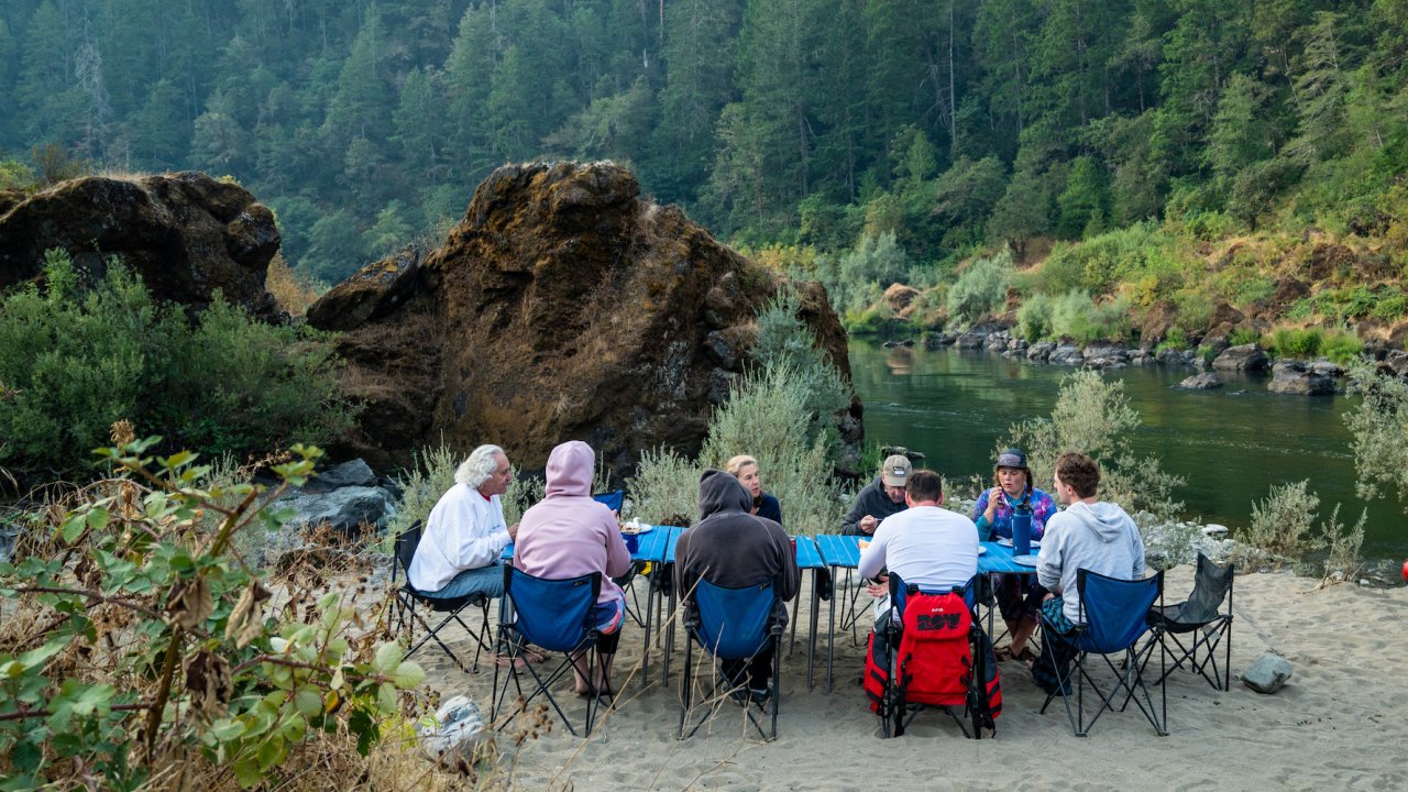 People sitting around blue tables in a beach while camping along a river in Oregon