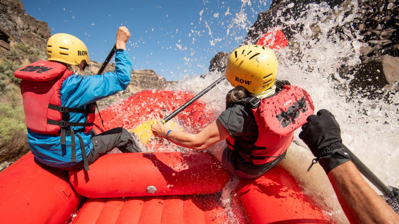 The back of two people wearing ROW Adventures life jackets as they get splashed while paddling on a red raft through a whitewater rapid