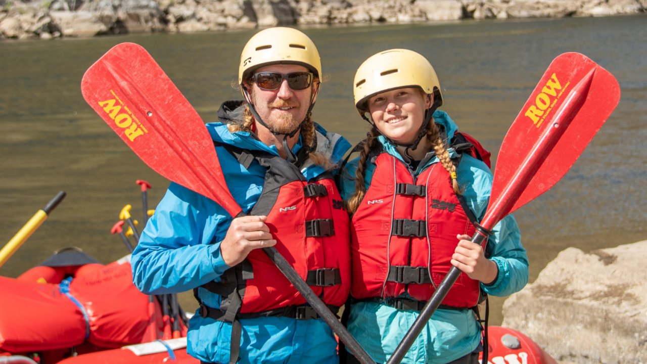 Two guests smiling while holding red ROW branded paddles and wearing yellow helmets about to start whitewater rafting