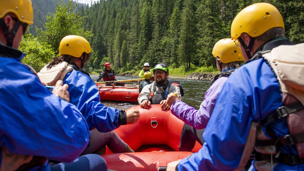 Passengers sitting on a red raft while their guide stands in the water at the bow of their boat giving a safety talk