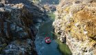 Aerial shot of a red raft floating down the Rogue River through a tight gorge with the right side in the sun and the left side of the gorge in the shade.