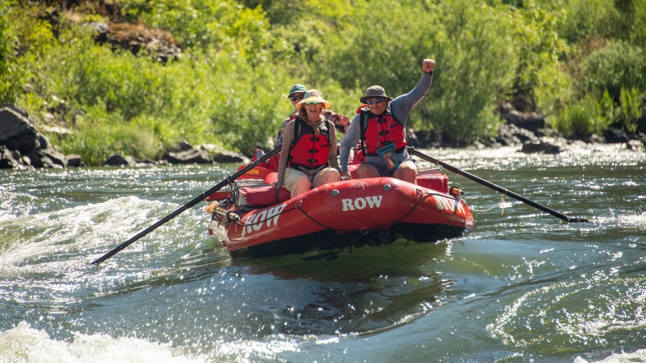 Group celebrating on a Rogue River rafting trip in Oregon, paddling through calm water surrounded by lush forest, perfect for rafting and camping adventures.