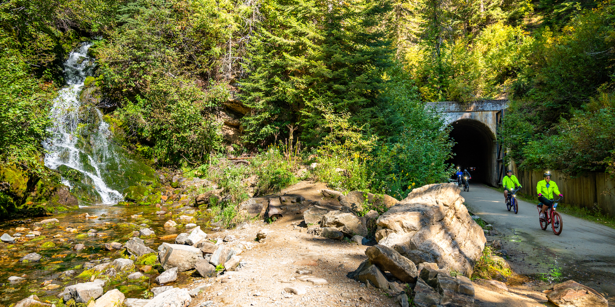 Bikers pedaling out of a tunnel on the Route of the Olympian next to a waterfall in North Idaho