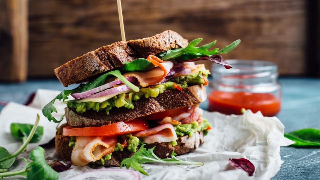 Big BLT sanswich on a white plate next to a red drink in a glass