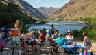 Campers sitting around square blue tables on a sandy beach along the Snake River in Idaho