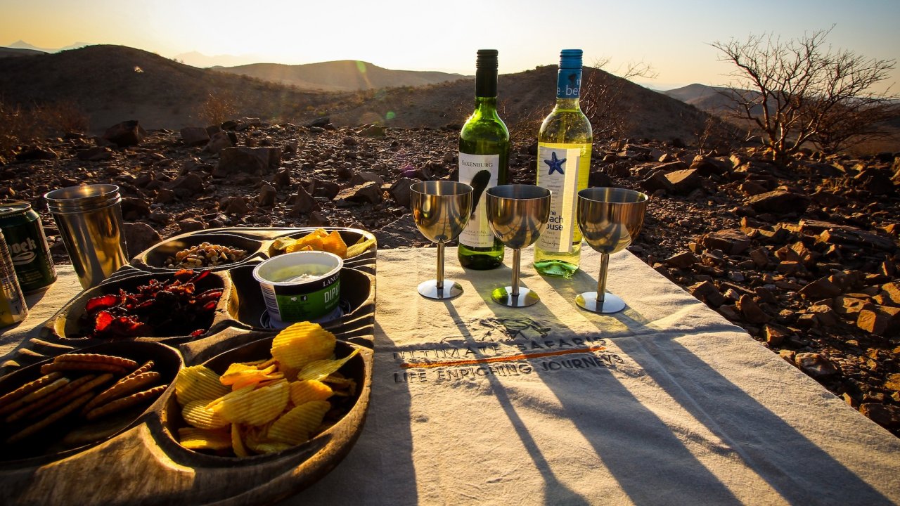Three metal wine glasses in front of two bottles of white wine next to a tray of chips and other light snacks overlooking the African desert at sunset