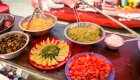 Ingredients for a taco salad bar laid out on a table under an umbrella on the beach 