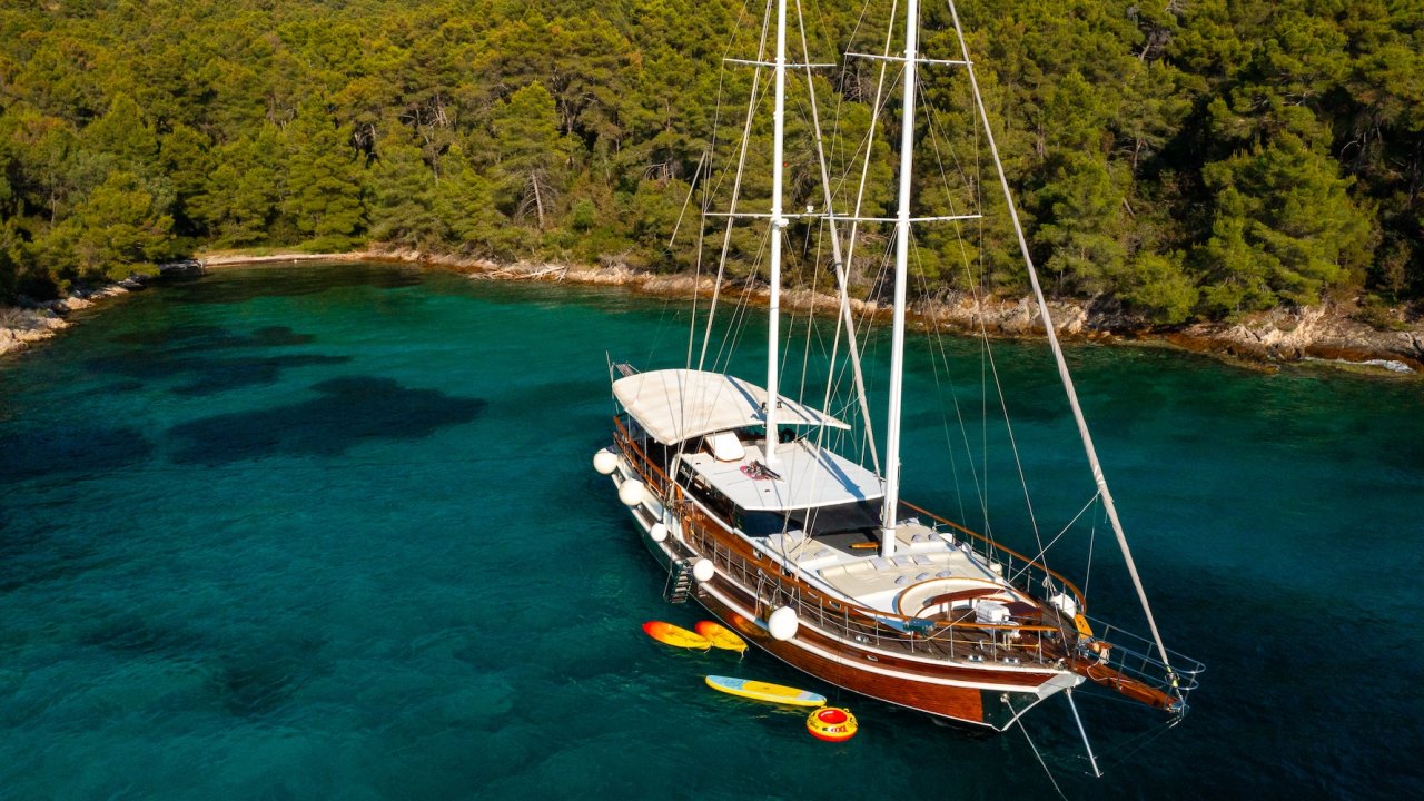 A sailing yacht anchored in a bay with paddle boards and inflatable water toys floating nearby.