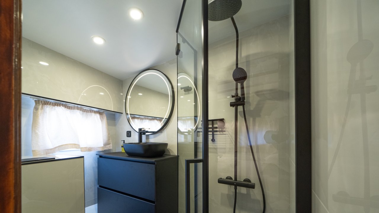 A yacht bathroom with a glass-enclosed rainfall shower, black vanity, and round mirror.